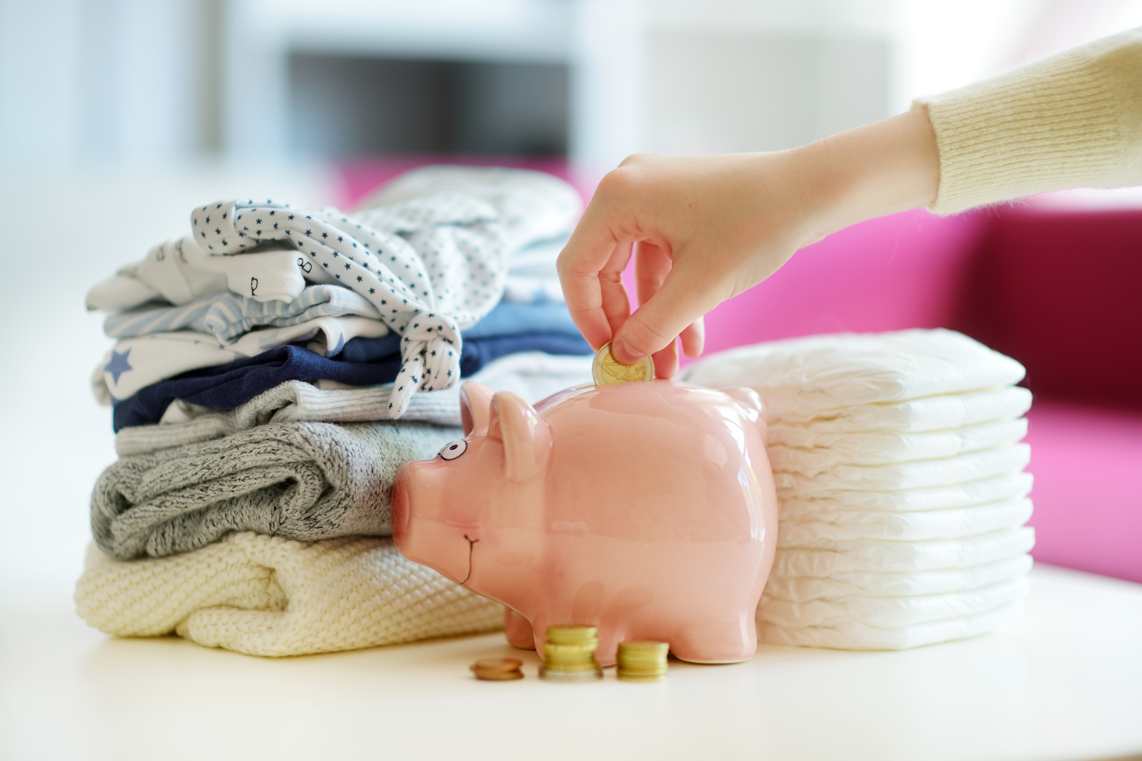 Hand Putting Coins in Piggy Bank with Baby Items on Table 